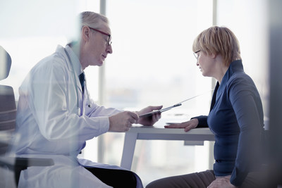 white haired man in glasses wearing a white coat seated next to a women in a blue shirt wearing glasses both looking at a document the man is holding
