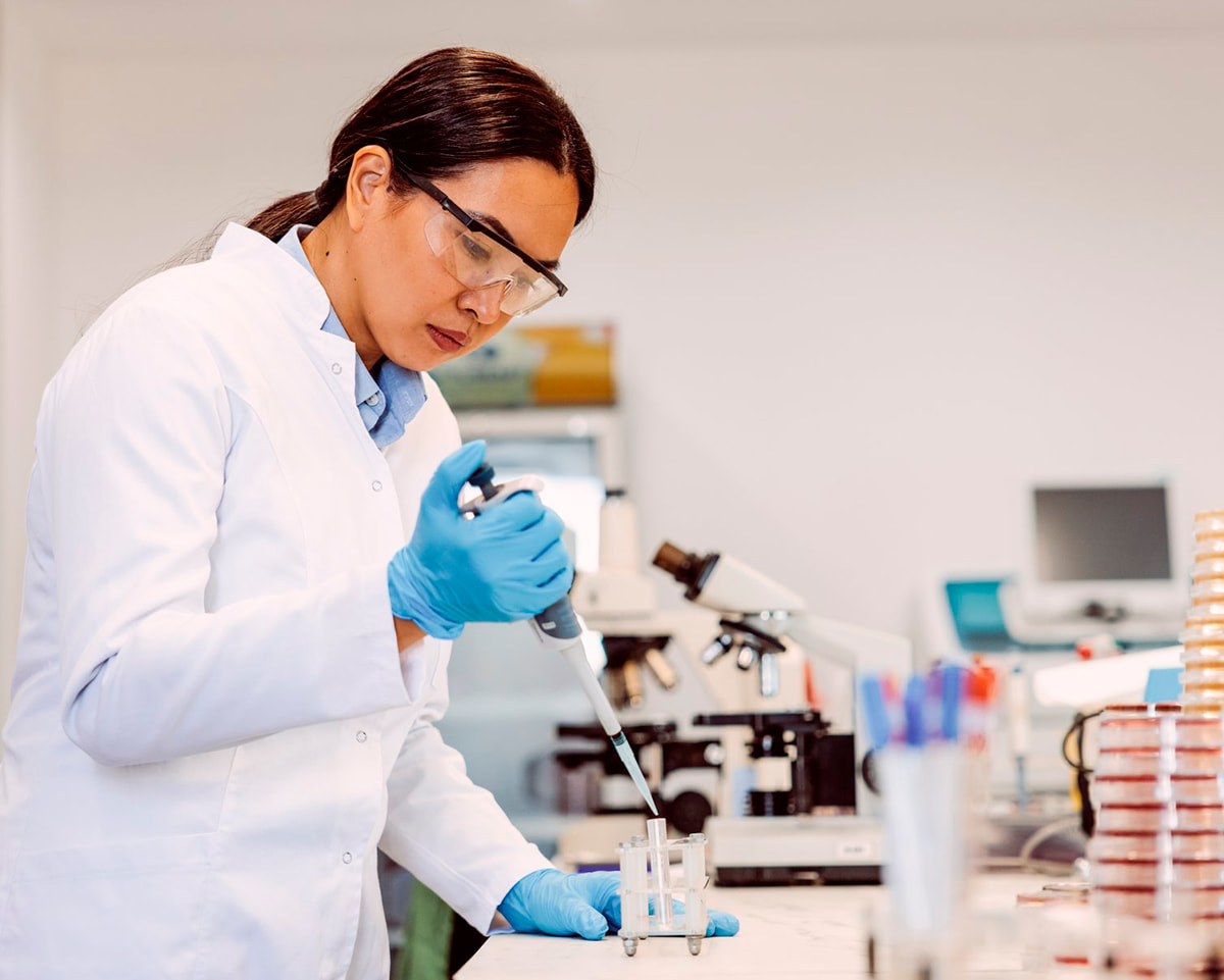 Female scientist using a single pipette; microscope and other lab equipment in the background on the lab bench.