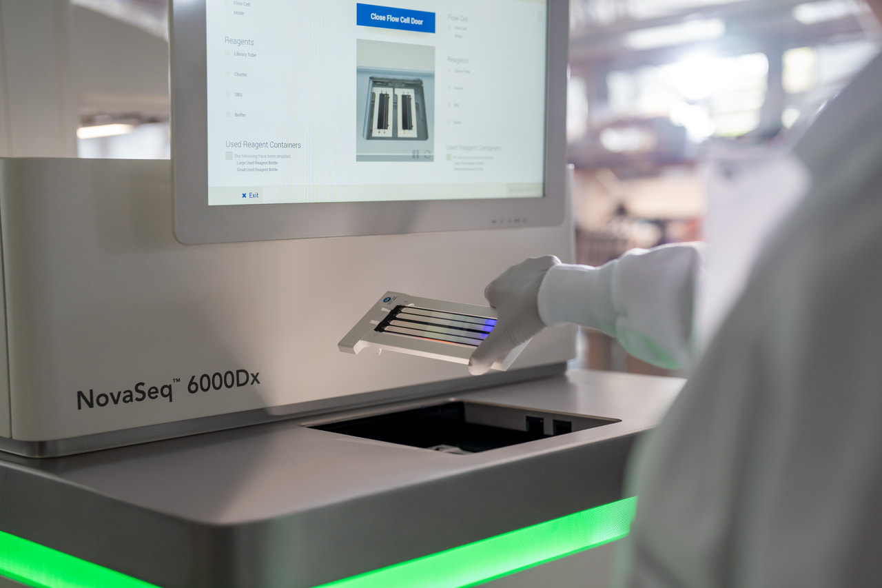 Close up, back view of a scientist inserting a flow cell in the flow cell compartment of a NovaSeq 6000dx; partial view of flow cell touch screen and green status bar visible.