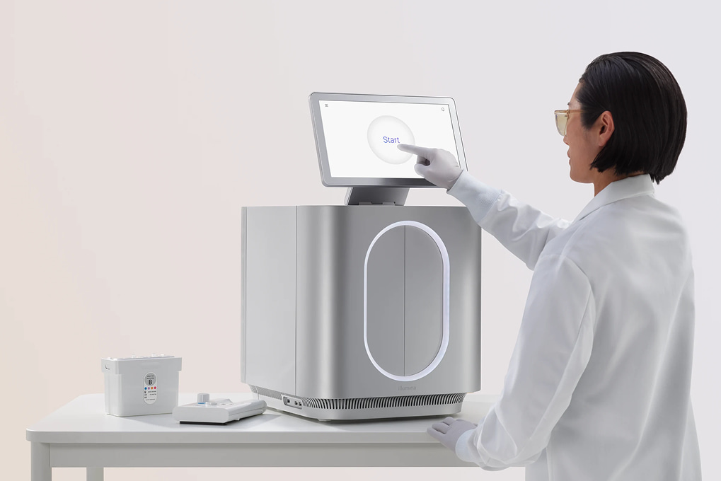 Side view of female scientist touching the start screen, instrument and wet and dry cartridges on lab bench, gradient background.