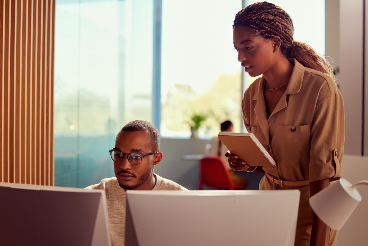 Male analyst, front view, sitting in front of computer screens with female standing behind discussing